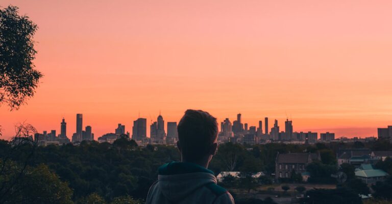 Man in Green Hoodie Looking at City Buildings during Sunset