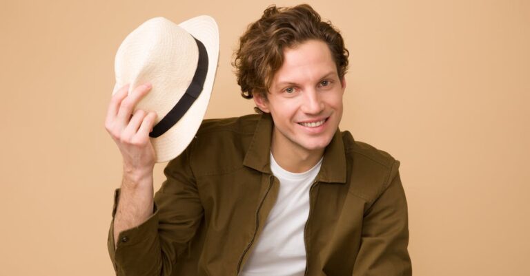 A cheerful man with curly hair tips his hat in a bright studio portrait.