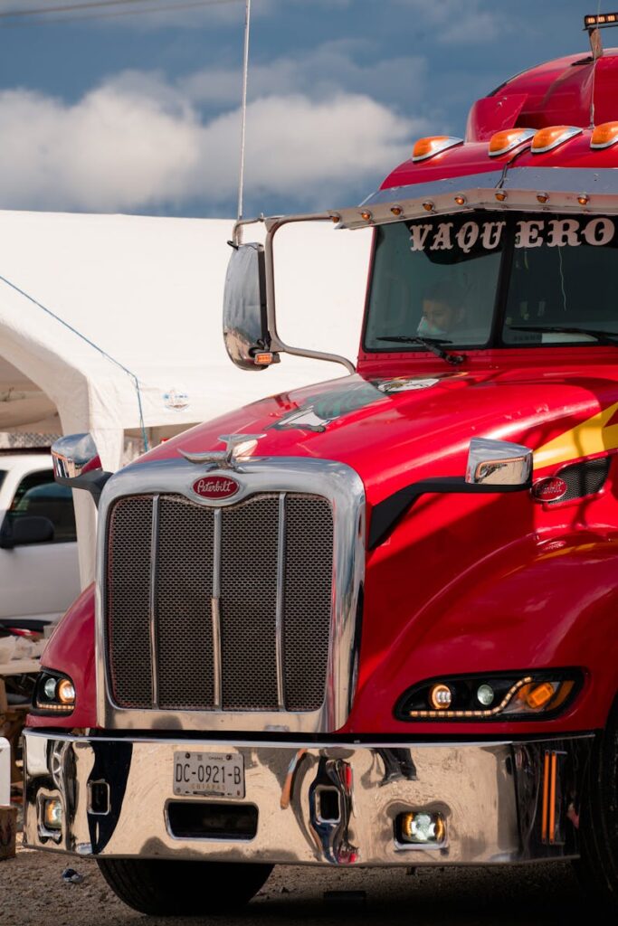 Close-up of a vibrant red semi-truck with chrome details at an outdoor event under cloudy sky.