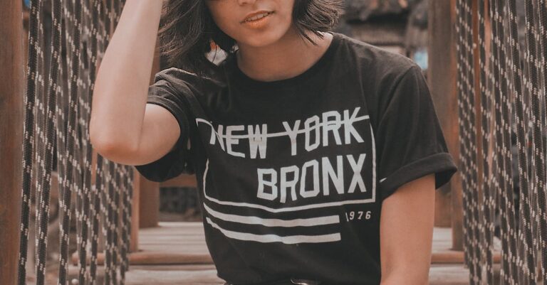 Confident woman in glasses wearing New York Bronx shirt sitting on a rustic wooden bridge outdoors.