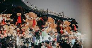 A Bustling Carnival Scene At Night Featuring Teddy Bear Prizes And A Lively Crowd.