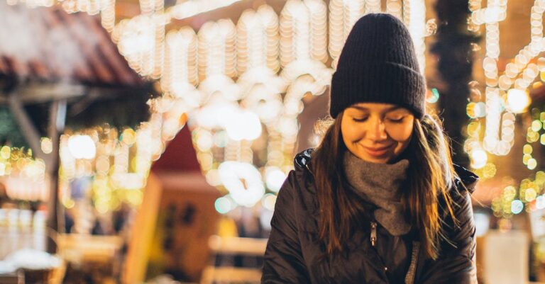 Happy young female in black warm jacket and hat looking down and smiling while sitting on wooden bench among Christmas decorations in night city