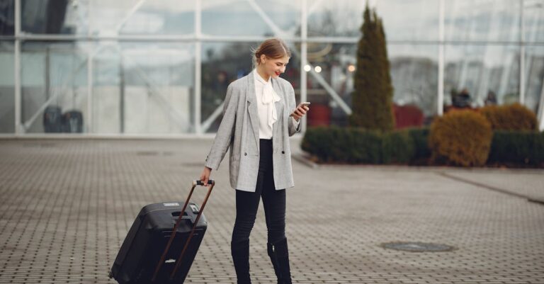 Confident businesswoman using smartphone while traveling with luggage outside an airport terminal.