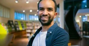 Smiling ethnic bearded student with toothy smile standing against bookshelves in library in college