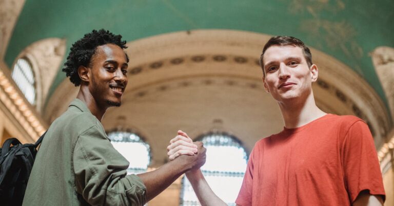 From below of positive young multiethnic male friends in casual clothes smiling and looking at camera while shaking hands in spacious aged arched building