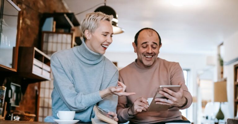 Cheerful adult diverse couple in casual clothes watching funny video on tablet and laughing while drinking coffee at table in apartment