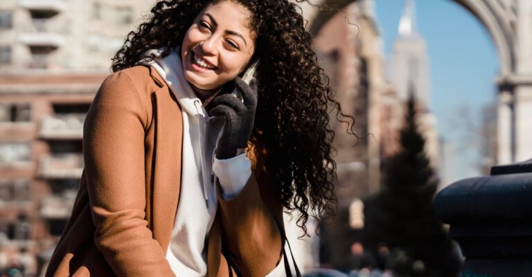 Young woman with curly hair happily chatting on phone in a city setting, enjoying a sunny day.