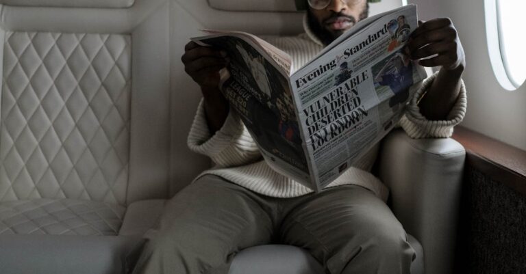 African American man reading newspaper and listening to music in airplane cabin with headphones.