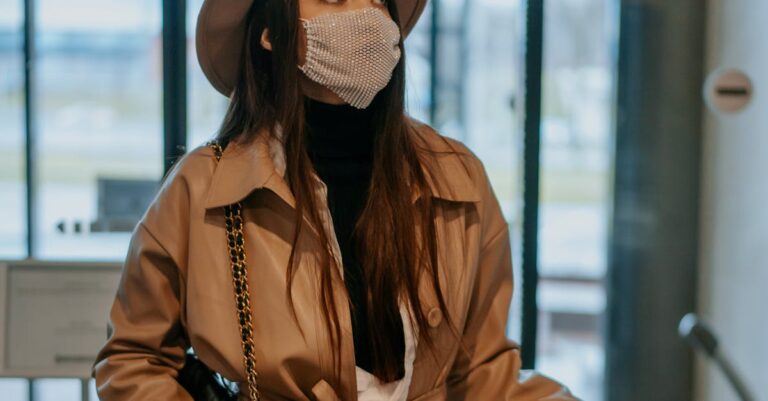 A fashionable woman in a brown coat and hat wearing a face mask inside an airport lobby.