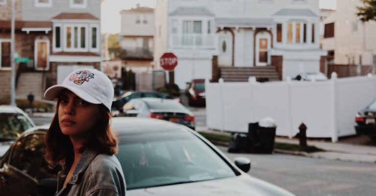 A young woman wearing a cap stands by a car in a residential New York neighborhood.