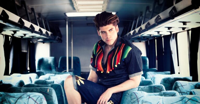 A stylish man poses confidently inside an empty bus wearing a colorful outfit.