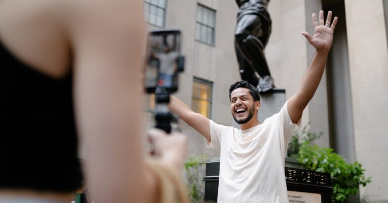 Smiling man raises arms joyfully near famous sculpture outdoors.