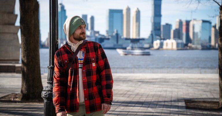 A man in casual winter attire stands with Manhattan skyline in the background.