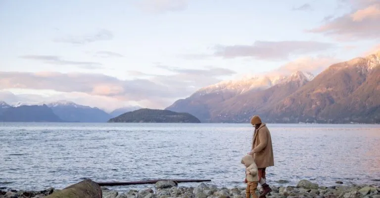 Side view of mother and little kid in casual warm outwear walking together along calm water on stony shore and holding hands