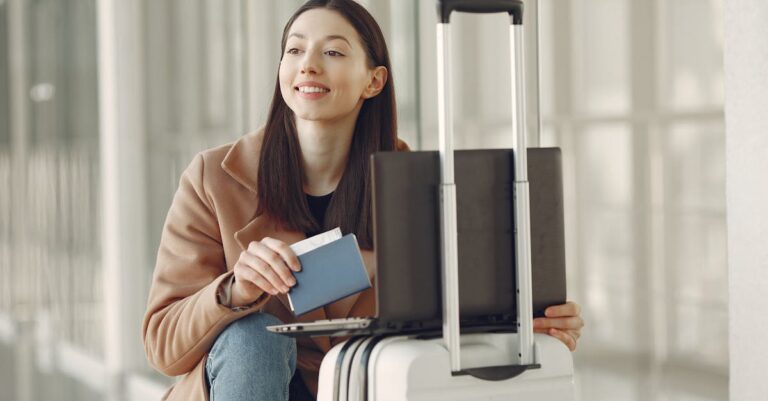 A woman sitting with her laptop and luggage, holding a passport, waiting in an airport terminal.