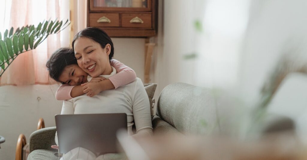 Excited cute Asian daughter bonding with smiling happy mother using netbook on cozy sofa in living room in daytime