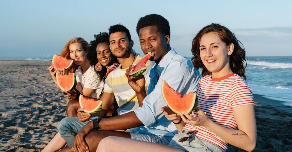 Young content multiracial friends enjoying sweet ripe watermelon slices on sandy sea shore while looking at camera