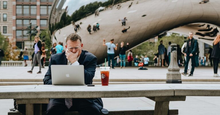 Concentrated businessman in suit touching face and sitting at table with laptop and takeaway drink against modern art sculpture Cloud Gate in Chicago in daytime