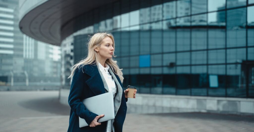 Confident businesswoman walking outside modern office building holding laptop and coffee.