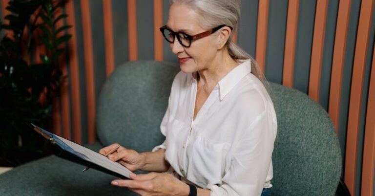 Elderly woman in glasses reviewing documents on clipboard, sitting indoors with a focused expression.