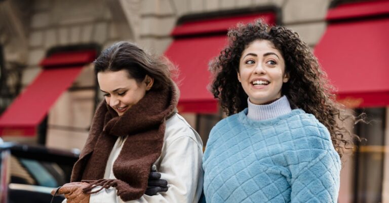 Cheerful trendy young multiethnic female millennials in stylish warm clothes smiling while strolling on city street near aged building