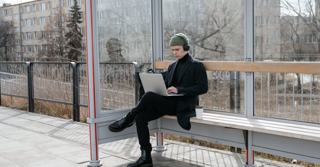 A young adult sits at a bus stop using a laptop, wearing headphones, during a sunny day.