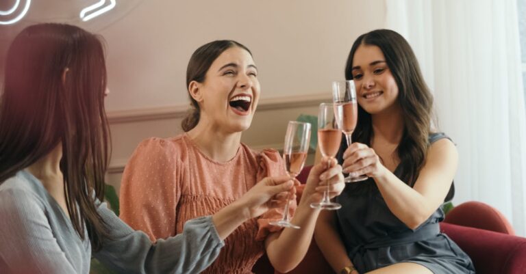 Three women laughing and clinking champagne glasses in a joyful indoor celebration.