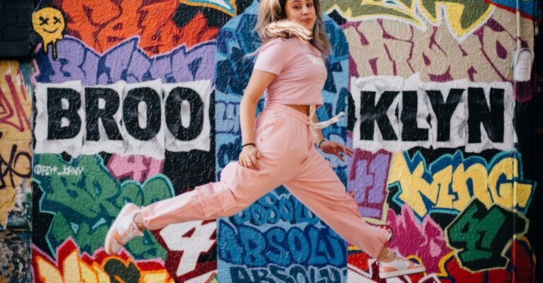 Energetic woman in pink jumpsuit jumping against a vibrant Brooklyn mural, capturing urban street art culture.