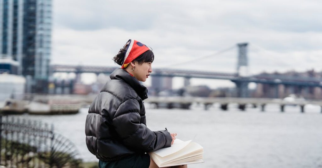 Side view of pensive Asian female taking notes in notebook and looking away while sitting on stone on embankment in city in daylight