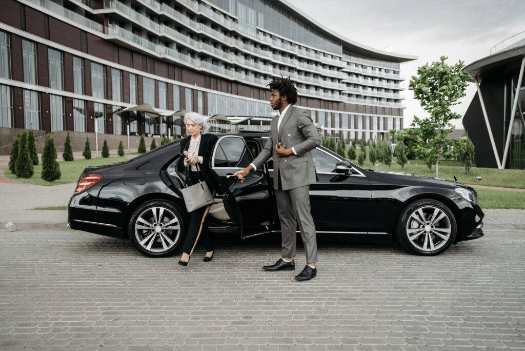 A Professional Chauffeur Assists A Businesswoman Exiting A Luxury Car In Front Of A Modern Building.