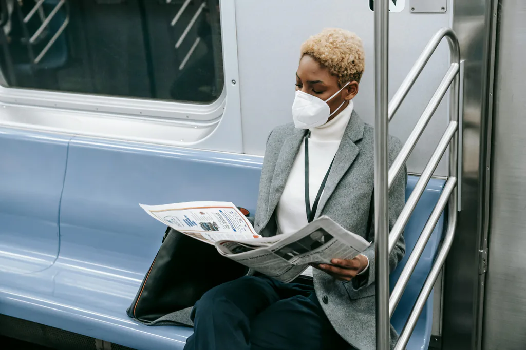 African American woman commuting on subway, reading newspaper and wearing a face mask.