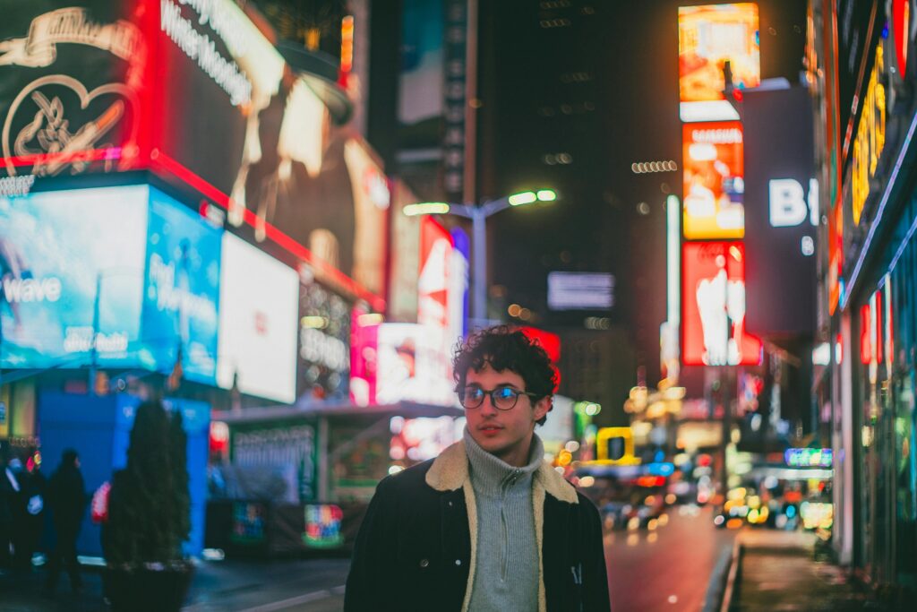 Young man in Times Square, New York City at night, surrounded by bright billboards and city lights.