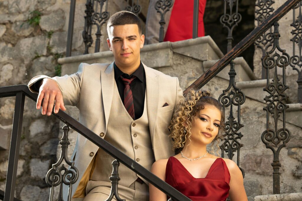 Stylish couple dressed formally posing on ornate staircase with stone walls.