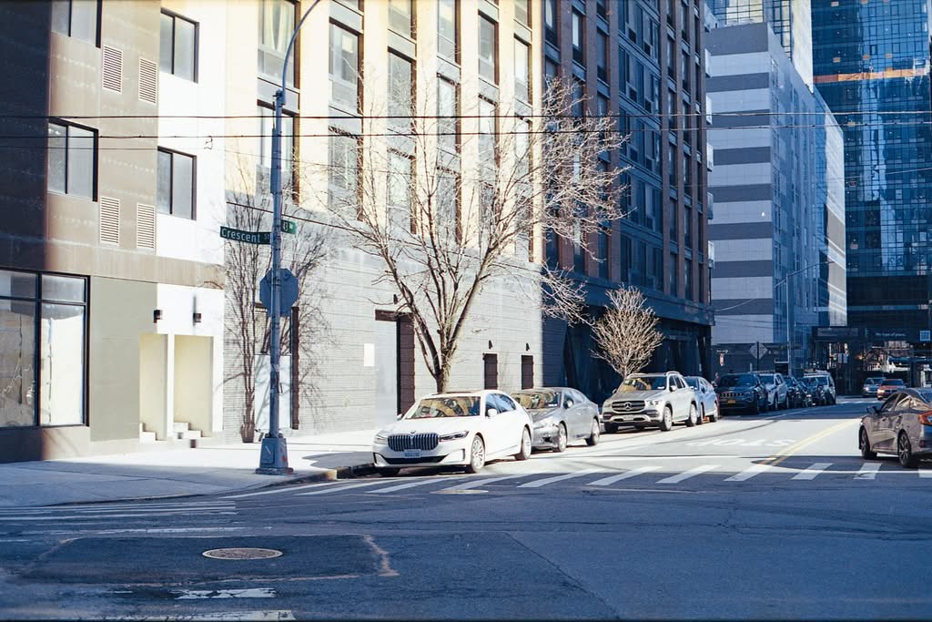 Sunlit street in New York City with parked cars and modern buildings.